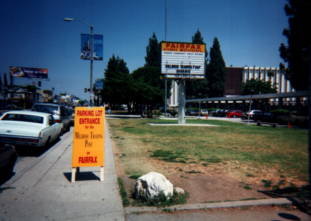 1997 film camera photo of the front (north) side of Fairfax HIgh School along Melrose Avenue. Sign reads Parking Lot Entrance for the Melrose Trading Post at Fairfax. Fairfax High School marquee reads, "Melrose Trading Post Sundays."